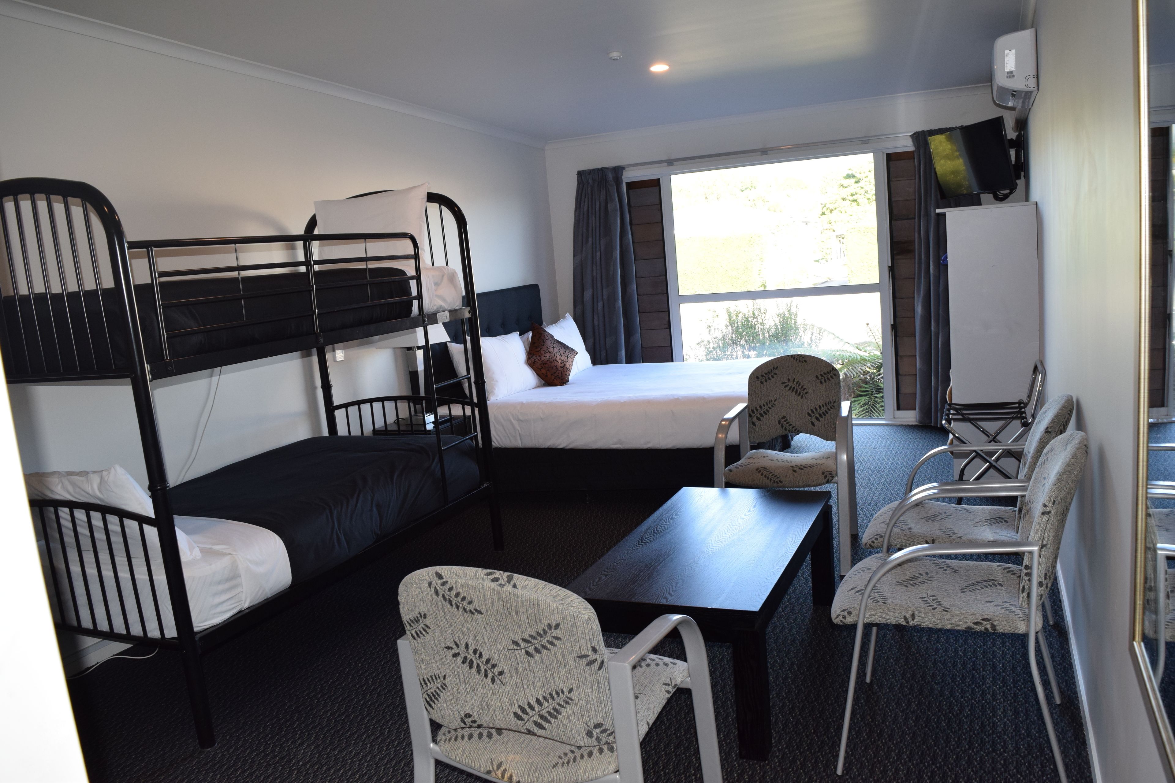 A dormitory room with bunk beds, a desk, chairs, a table, and a window with curtains.