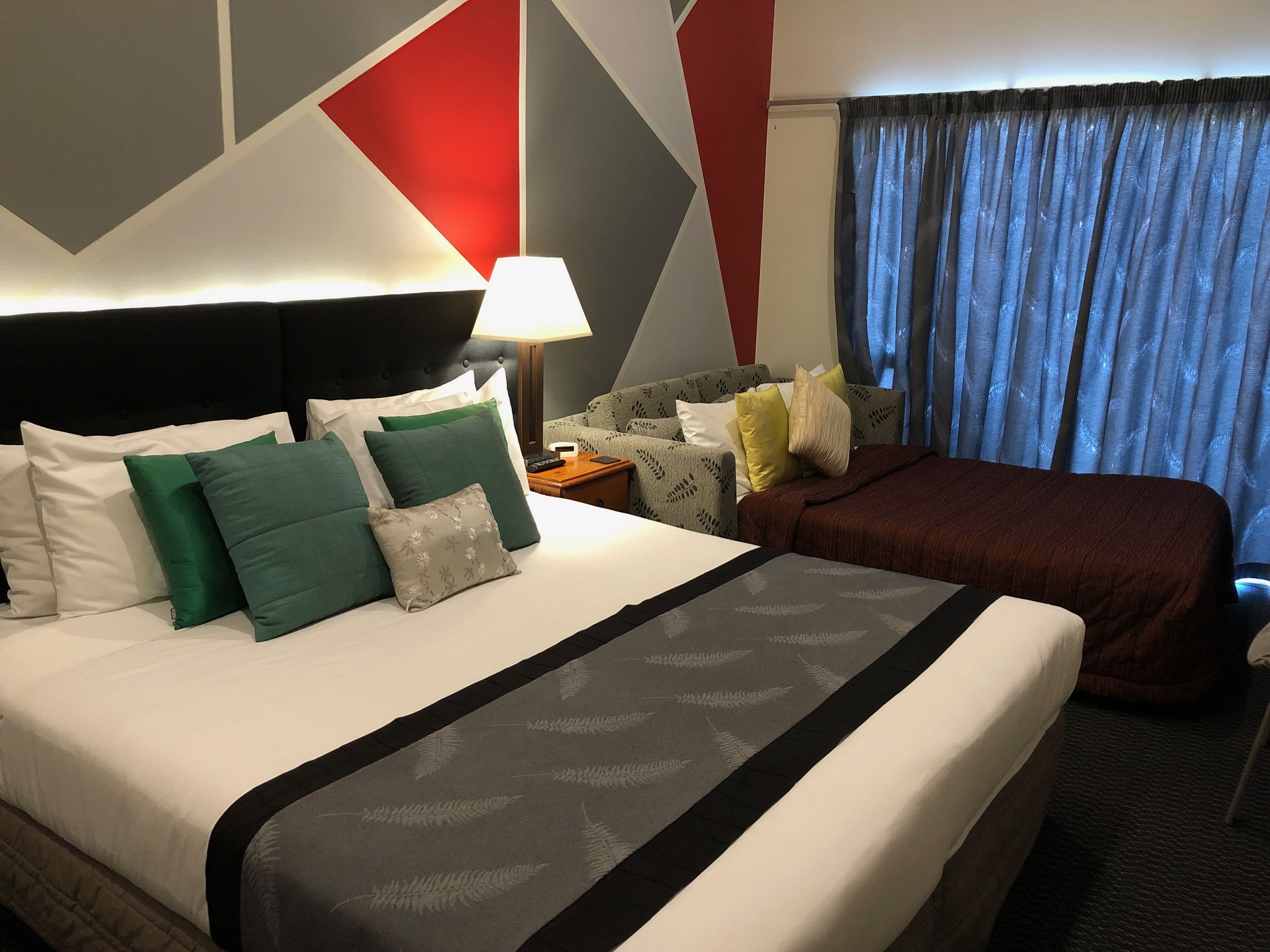 A hotel room with a large bed, a nightstand, a lamp, a sofa, and a view of the outside through curtains.