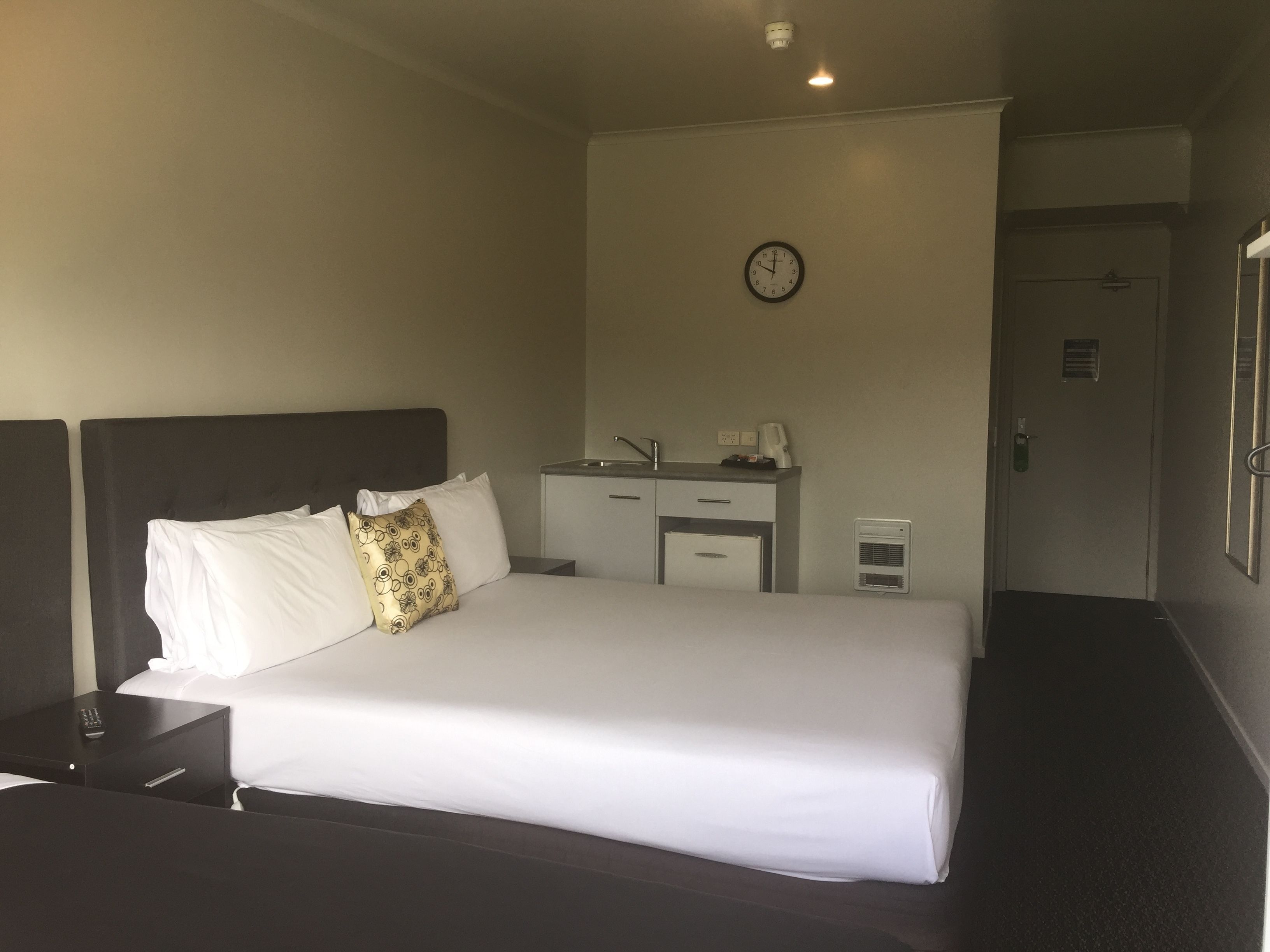 A hotel room with a bed, a nightstand, a desk, a chair, a clock, and a door.