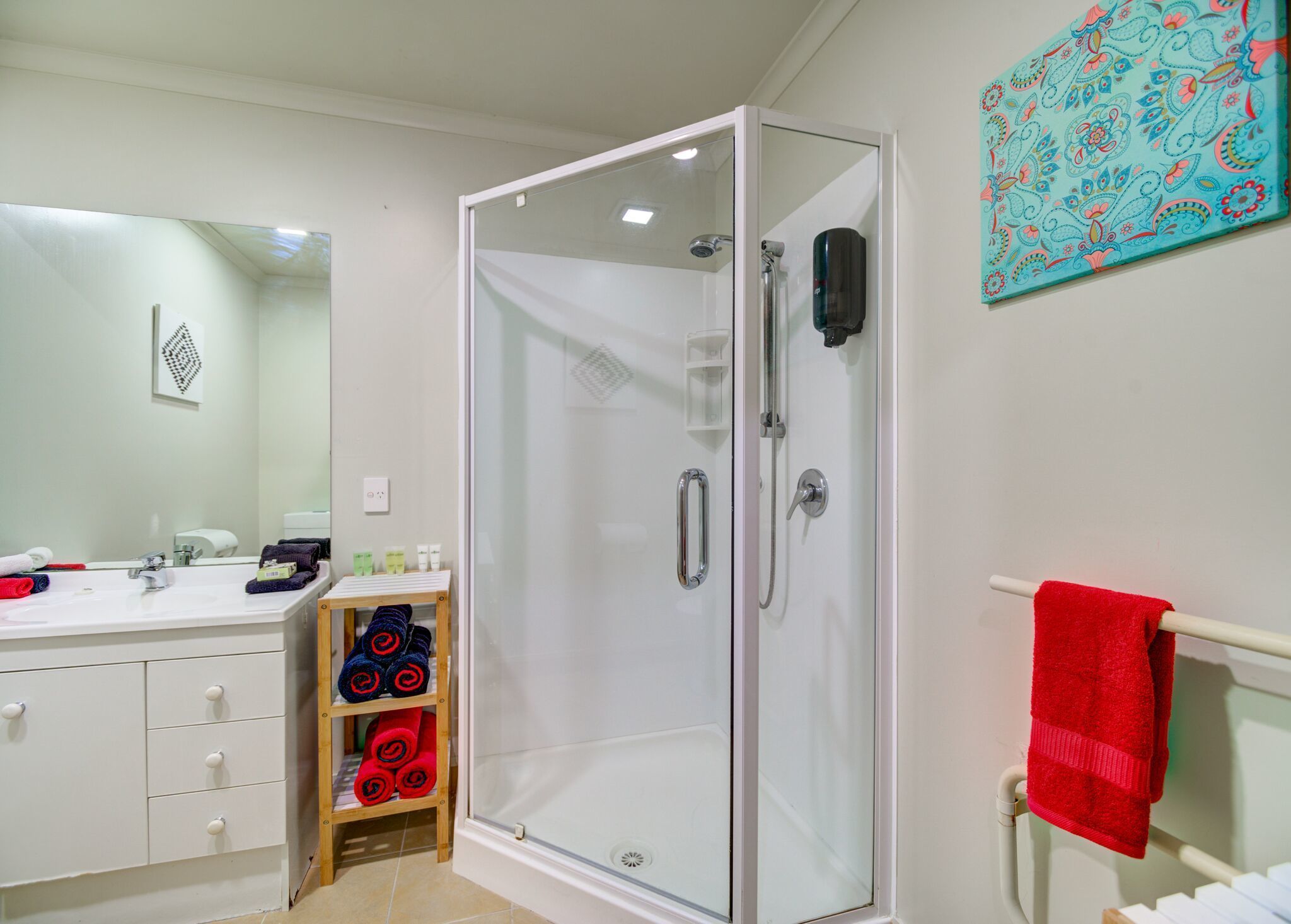 Un baño moderno con ducha de vidrio, un lavabo blanco con espejo y un perchero con una toalla roja.