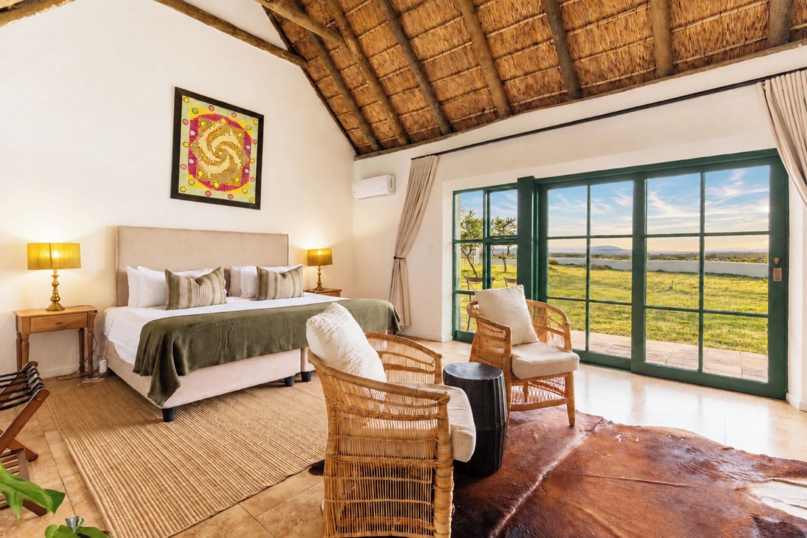 A bedroom with a large bed, two bedside lamps, a view of a grassy landscape through a large window, and a woven rug on the floor.