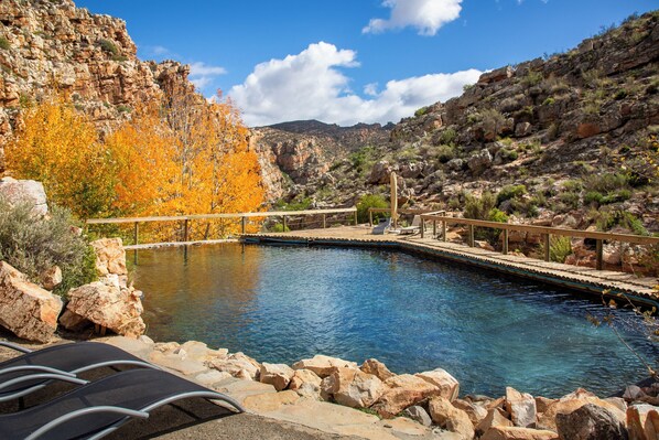 Zona de piscina con terraza de madera, sillas de descanso y paisaje rocoso al fondo.