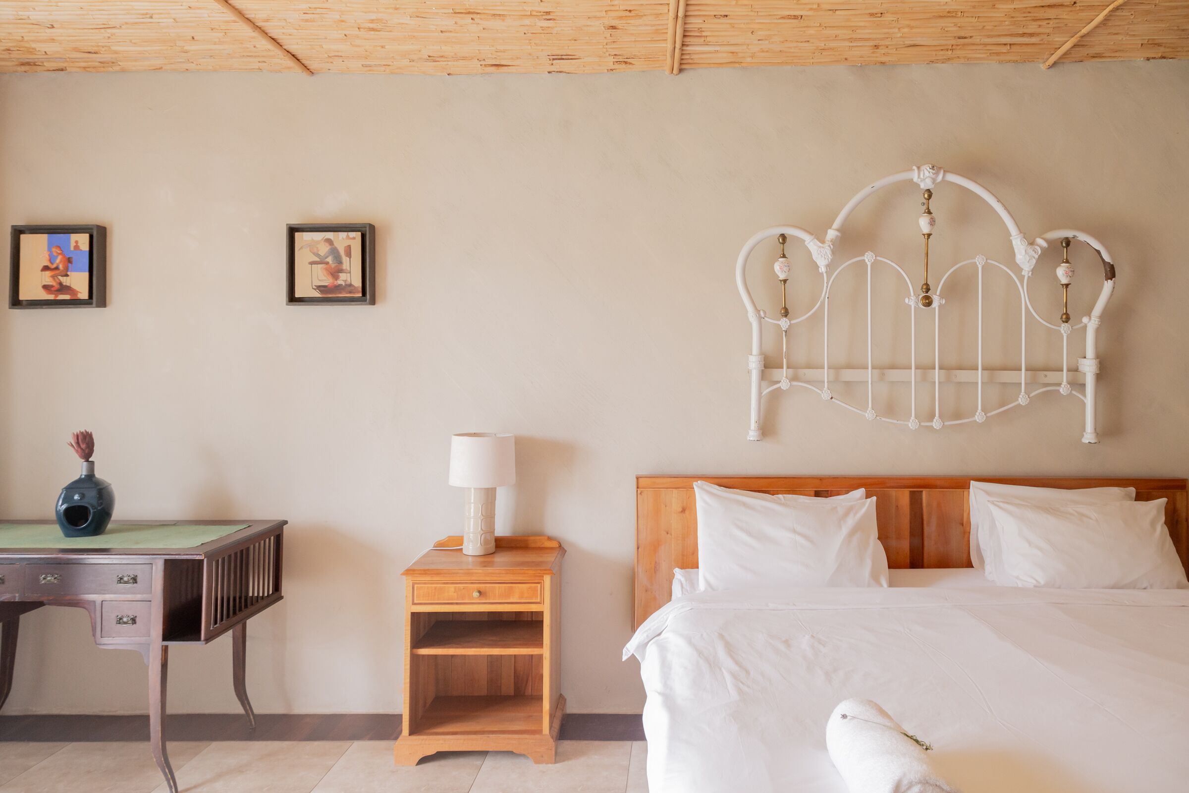 A neatly arranged bedroom with a wooden headboard, a bedside table with a lamp, and two framed pictures on the wall.