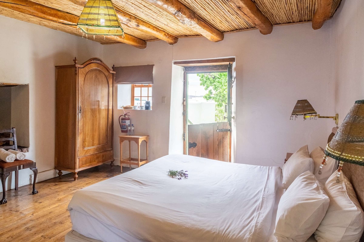 A bedroom with a wooden wardrobe, a bed with white linens, a wooden chair, a small table, and a window with a view of greenery.