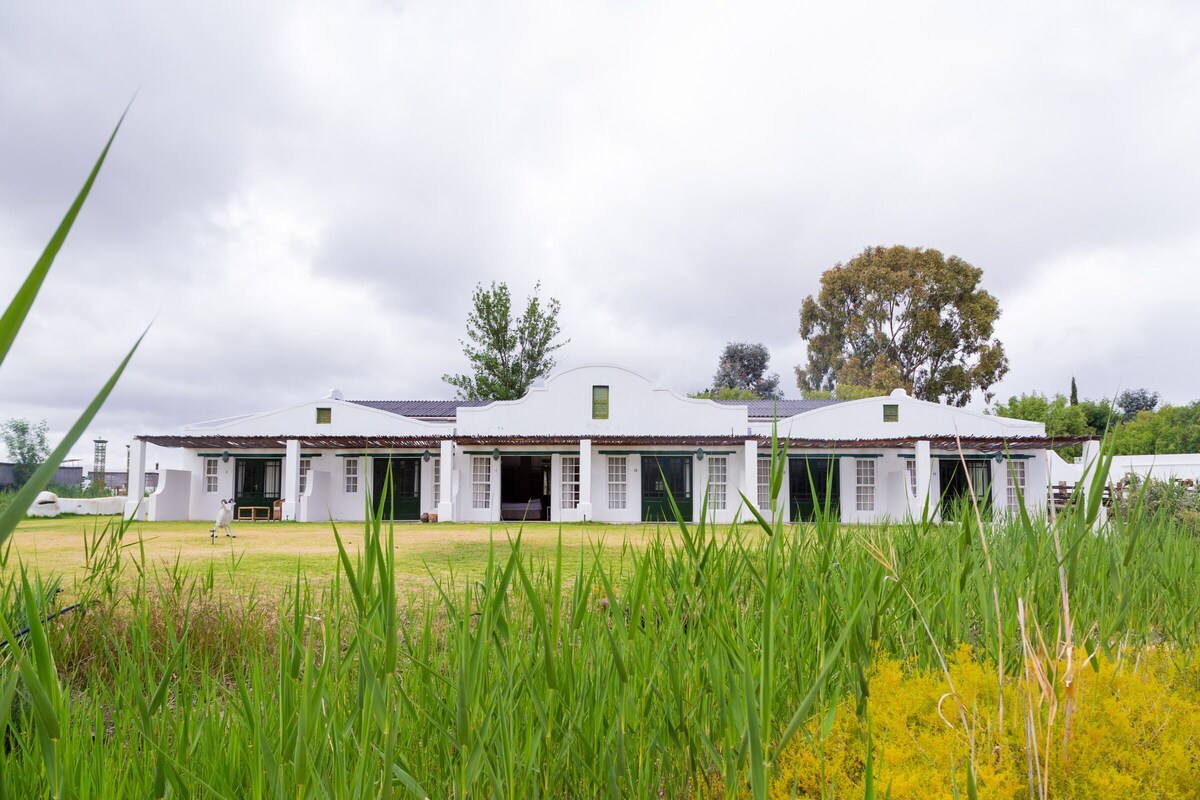 A white building with a flat roof and multiple doors, surrounded by tall grass and a tree.