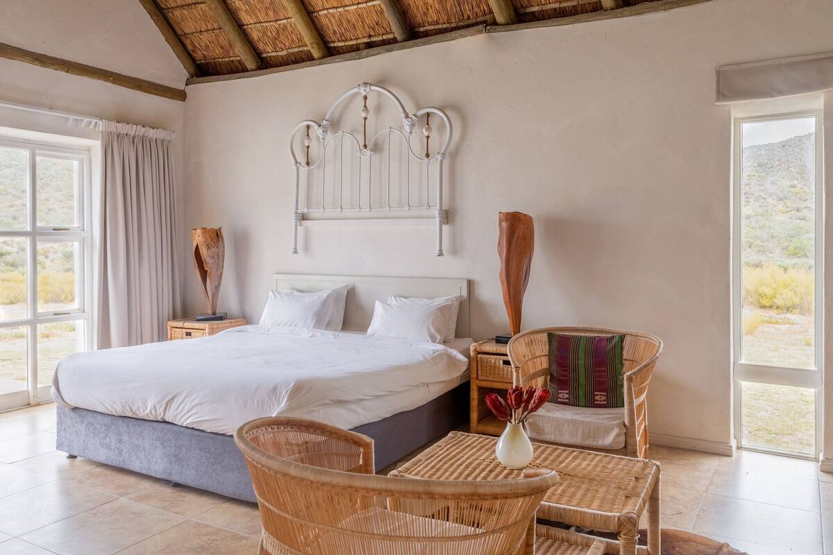 Een slaapkamer met een groot bed, een hoofdeinde, een nachtkastje, een rieten stoel, een kleine tafel en een raam met uitzicht op de omgeving.