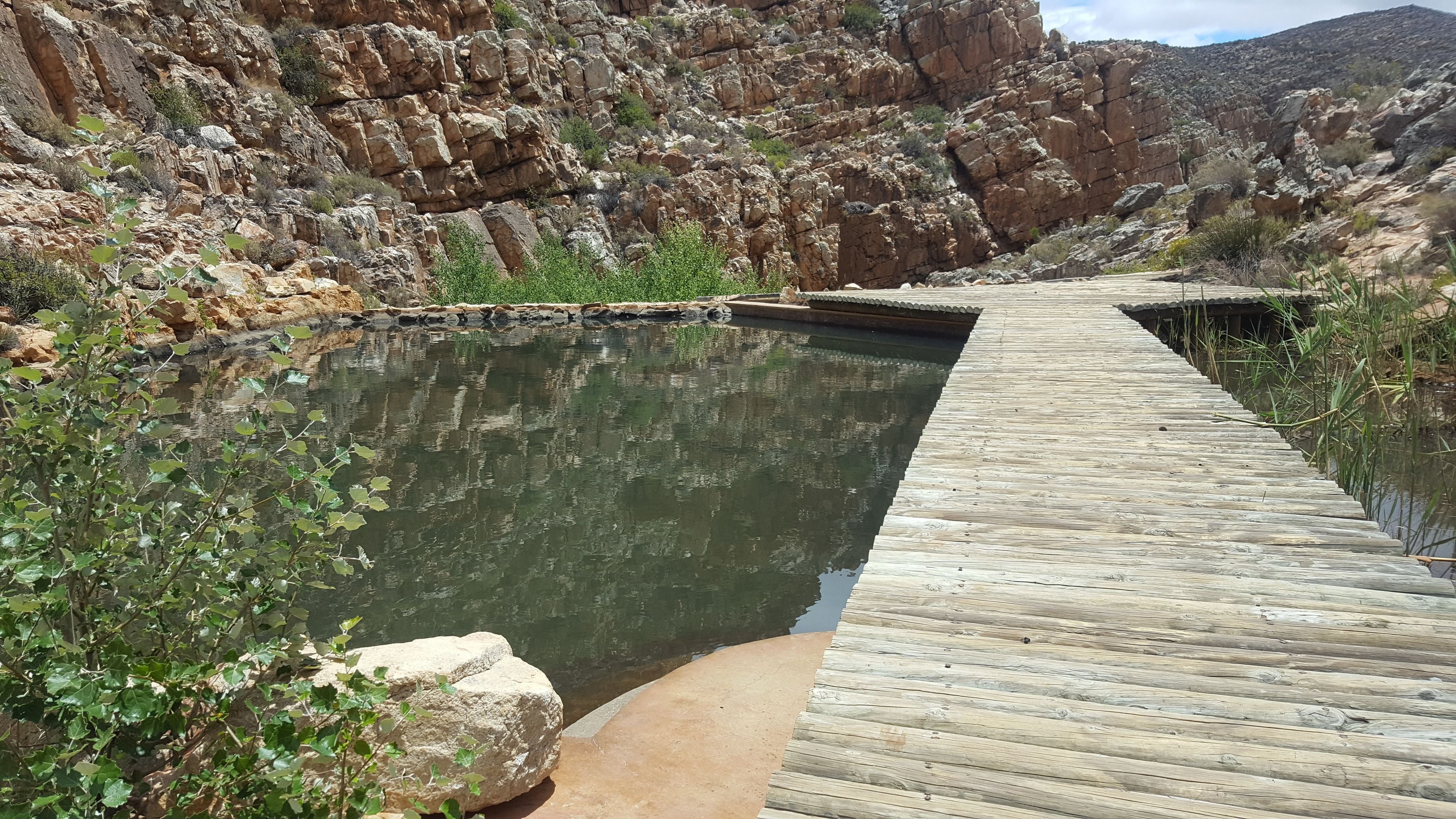 Eine Holzterrasse führt zu einem kleinen Pool, umgeben von felsigem Gelände und vereinzelter Vegetation.