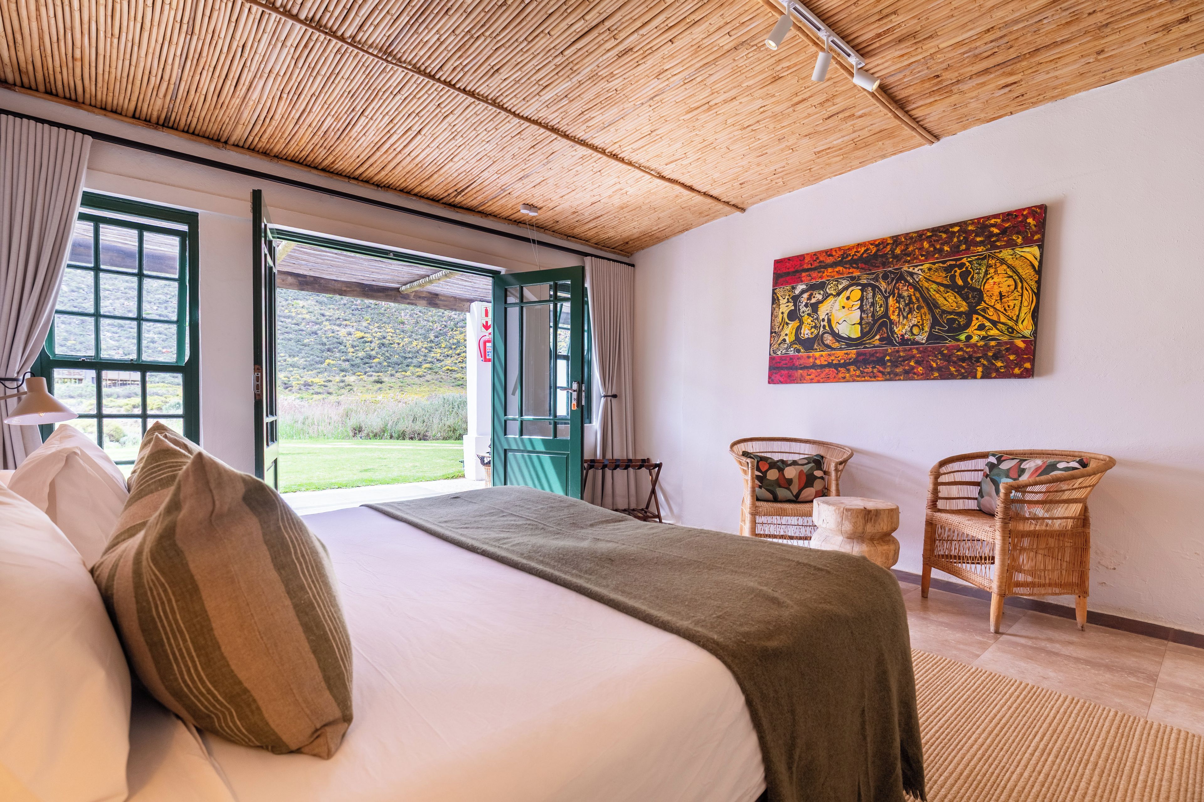 A bedroom with a bed, two chairs, a painting, and a view of the outdoors.