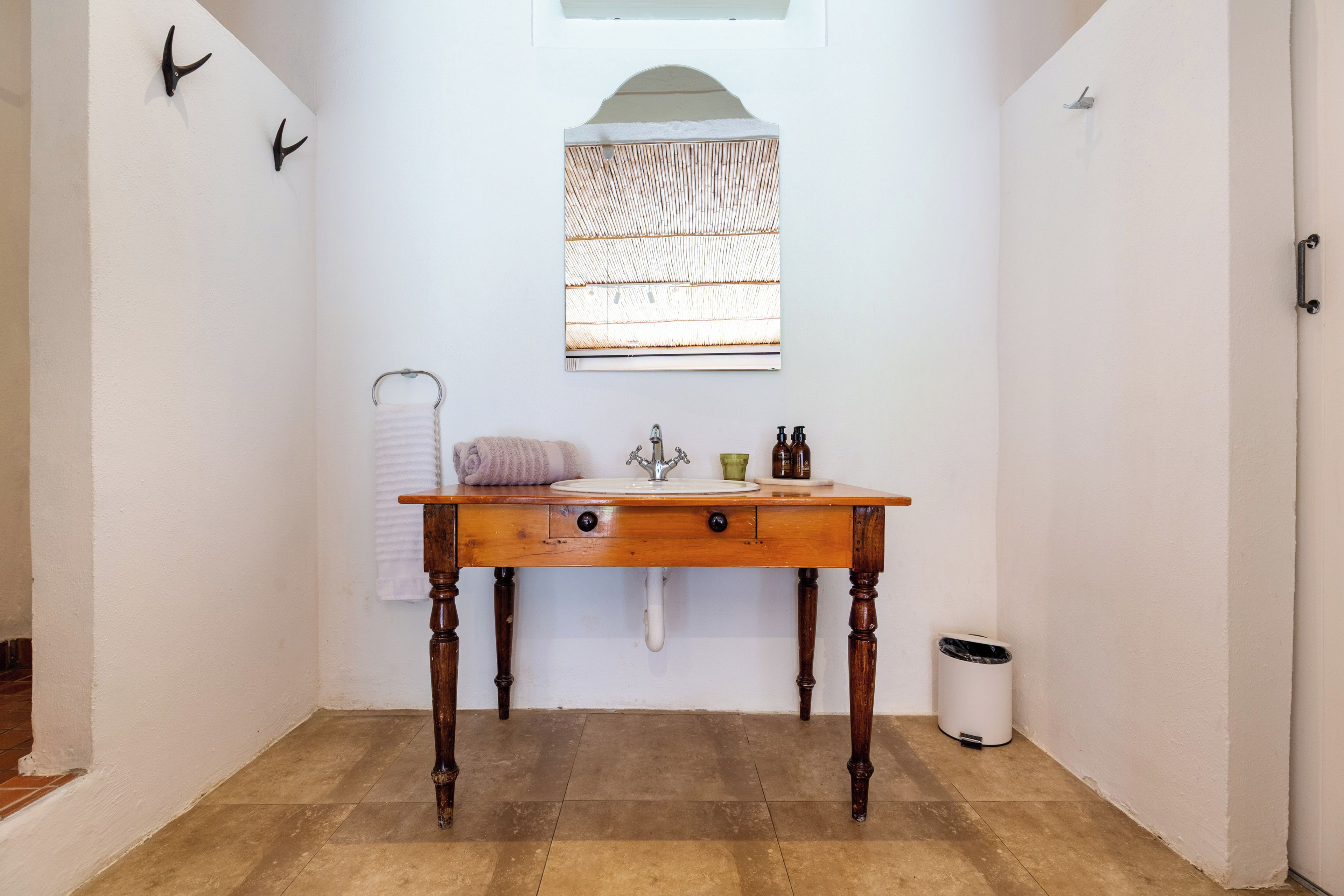 Un baño con tocador de madera, toallero, ventana con persiana de bambú y cubo de basura.