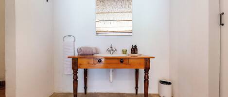 Un baño con tocador de madera, toallero, ventana con persiana de bambú y cubo de basura.