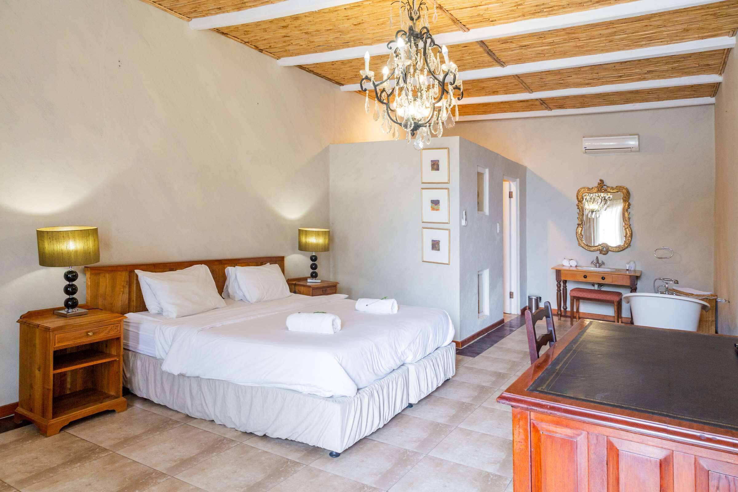 A hotel room with a large bed, wooden headboard, two bedside tables with lamps, a chandelier, a vanity with a mirror, and a bathtub.