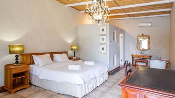 A hotel room with a large bed, wooden headboard, two bedside tables with lamps, a chandelier, a vanity with a mirror, and a bathtub.