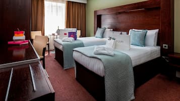 Habitación de hotel con dos camas, un escritorio y una silla. Se observan almohadas decorativas y una bolsa de regalo J'adore sobre las camas.