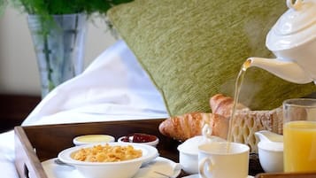 Bandeja de café da manhã com croissants, um bule servindo chá, uma xícara de café, uma tigela de cereal e um copo de suco de laranja sobre uma cama, com um travesseiro e um vaso de flores ao fundo.
