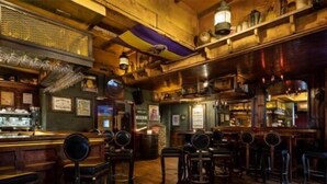 Eine Bar mit Holzböden und Barhocker, einer Theke mit Registrierkasse und Regalen mit Flaschen und Gläsern.