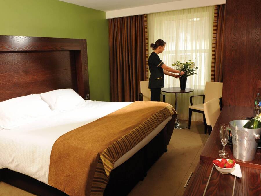 A hotel room with a large bed, a wooden headboard, a dining table with a vase of flowers, a bottle of champagne, and a bowl of strawberries.