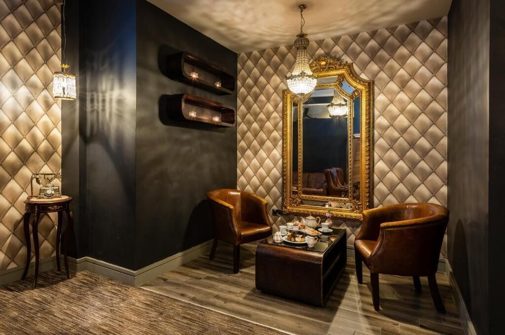 A room with a tufted wall, a large mirror, two leather chairs, a small table with tea set, and a hanging chandelier.