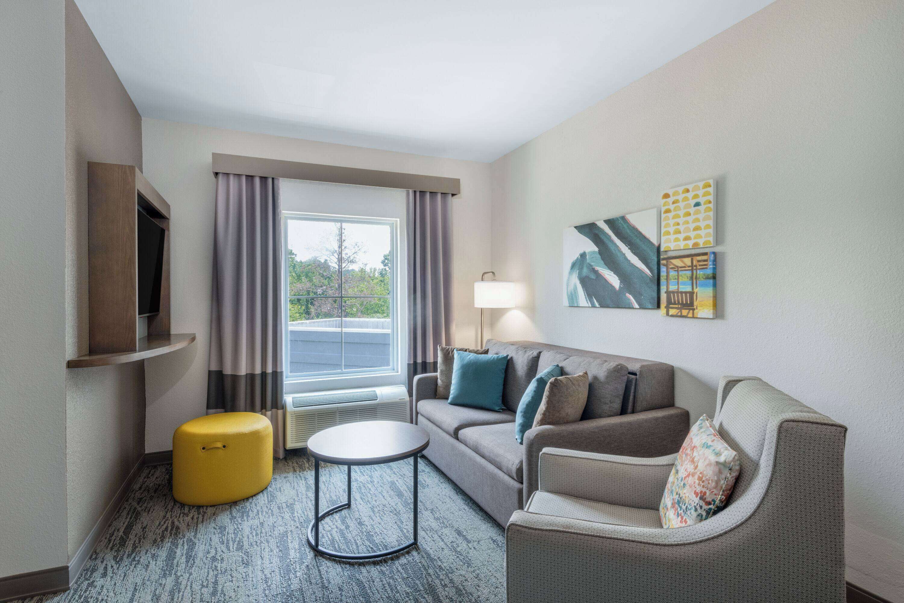 A hotel room with a sofa, armchair, ottoman, and a small round table. There is a window with curtains, a flat-screen TV mounted on the wall, and a lamp on the side table.