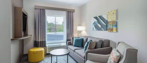 A hotel room with a sofa, armchair, ottoman, and a small round table. There is a window with curtains, a flat-screen TV mounted on the wall, and a lamp on the side table.