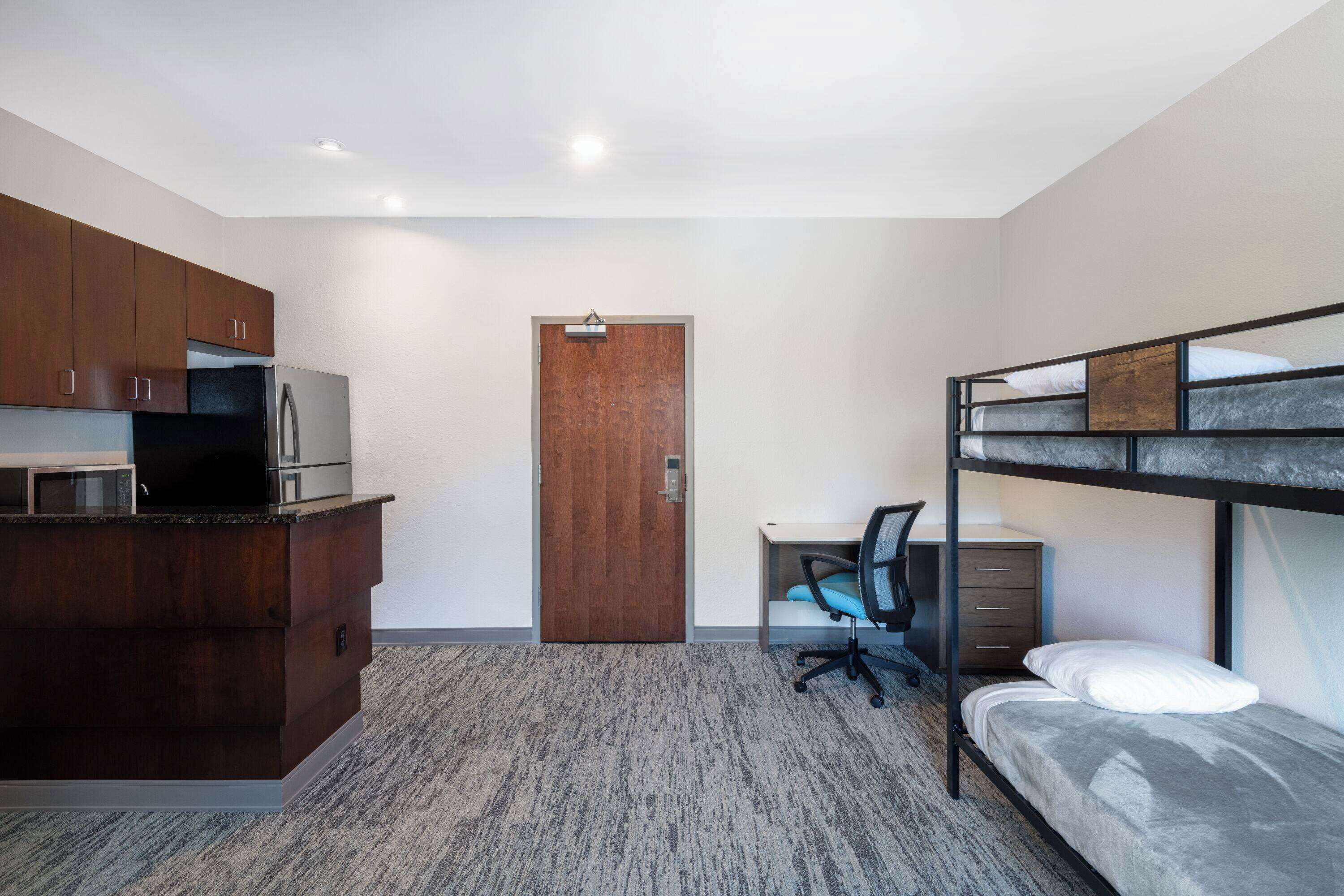 A compact room with a bunk bed, a kitchenette with a refrigerator and microwave, a wooden door, and a desk with a chair.