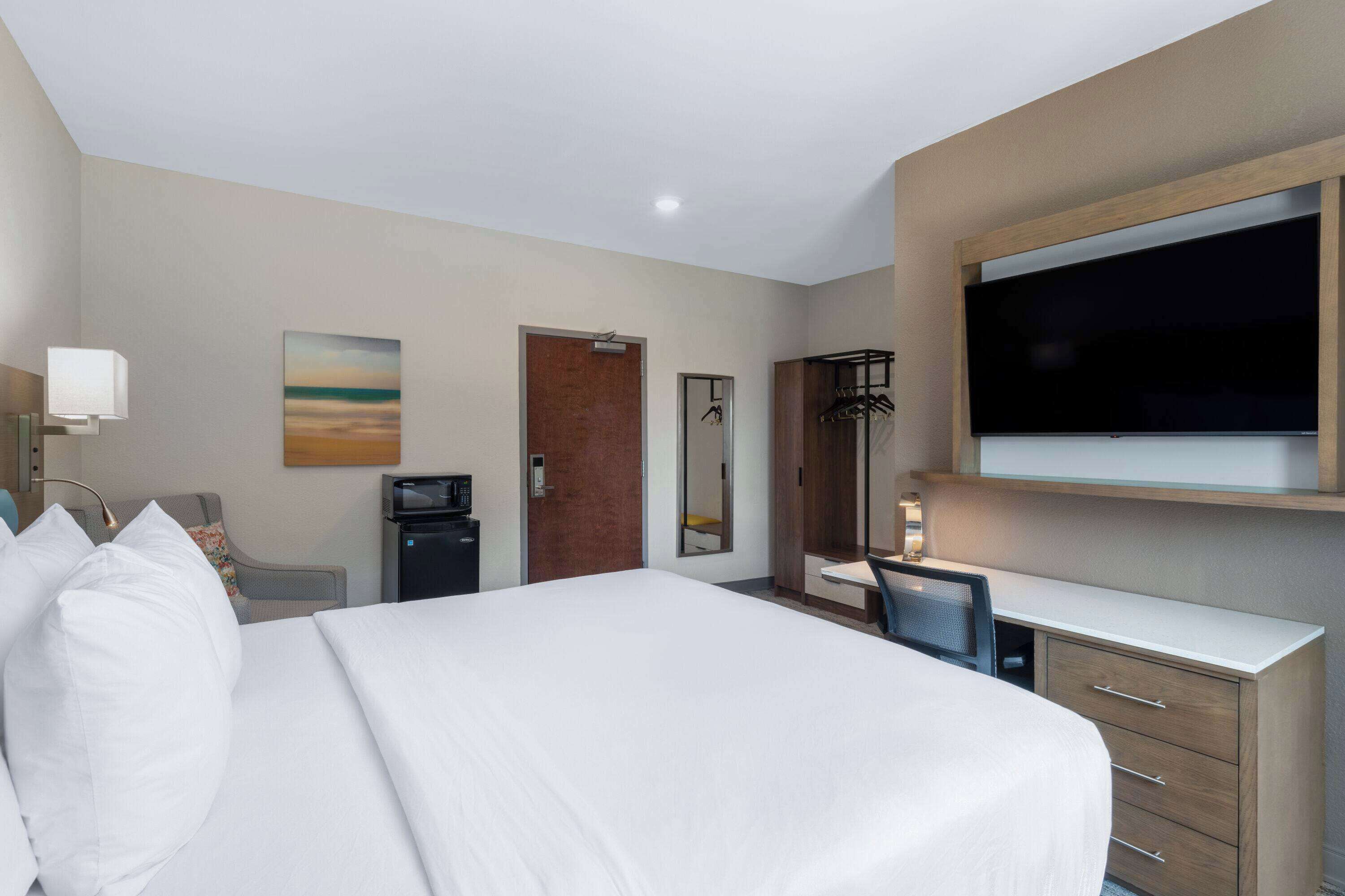 A hotel room with a large bed, a desk with a chair, a television, a mirror, and a door.