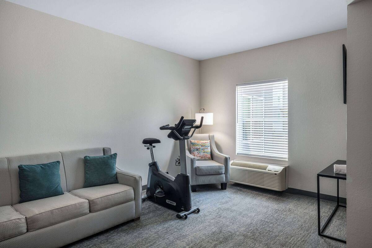Un salon comprenant un canapé, un vélo d’exercice, un fauteuil, une lampe et une fenêtre avec des stores.