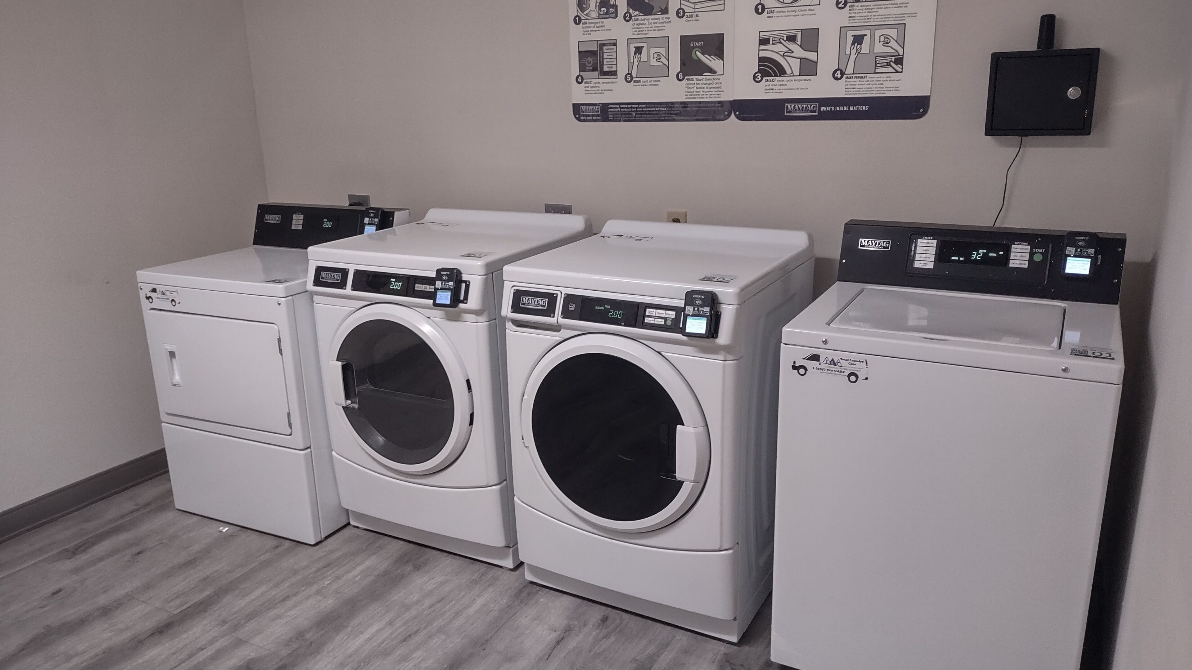 A laundry room with three washing machines and a dryer, a control panel with instructions, and a wall-mounted electrical outlet.