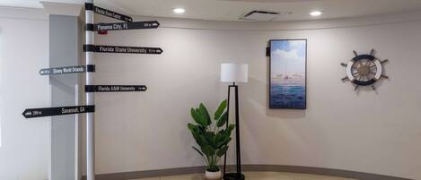 A lobby with a directional signpost, a potted plant, a floor lamp, a painting, and a ship's wheel decoration.