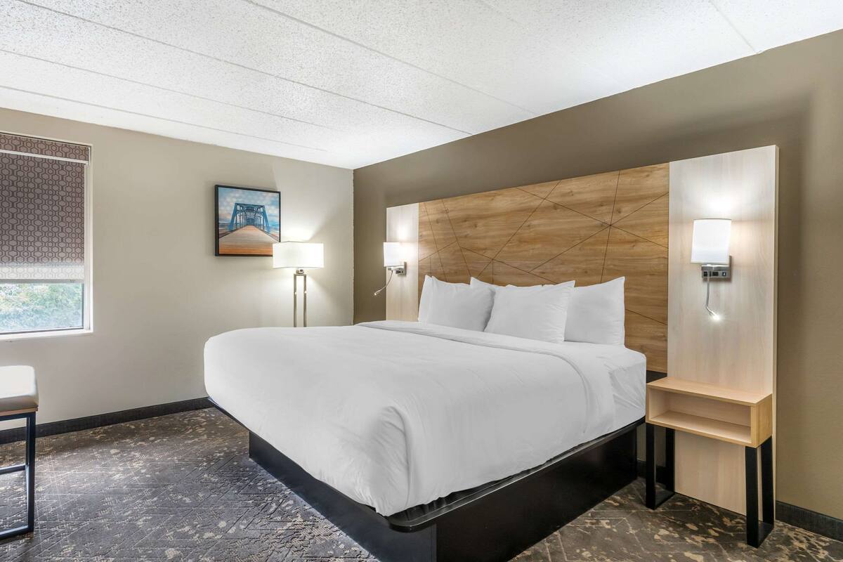 A modern hotel room with a large bed, a wooden headboard, two bedside lamps, a small table, and a window with a view of greenery.
