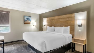 A modern hotel room with a large bed, a wooden headboard, two bedside lamps, a small table, and a window with a view of greenery.