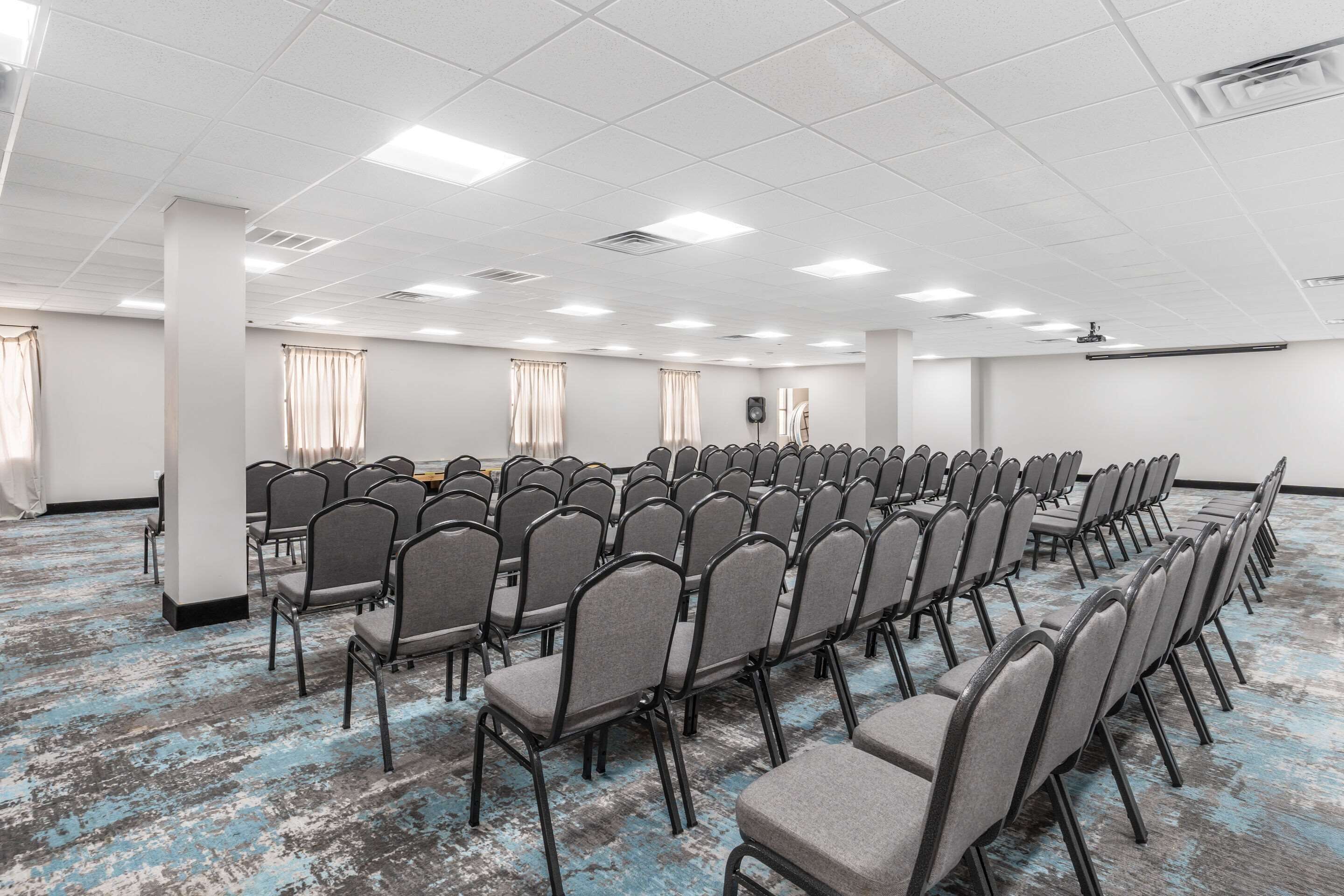 Une grande salle de conférence avec des rangées de chaises, un plafond blanc éclairé par des luminaires et une moquette bleue.