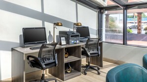 Un moderno ufficio con due scrivanie, ciascuna dotata di un computer, una stampante e una lampada. Sono presenti due sedie, una nera e una verde acqua. La stanza ha un'ampia finestra con vista su un parcheggio e un edificio con un cartello.