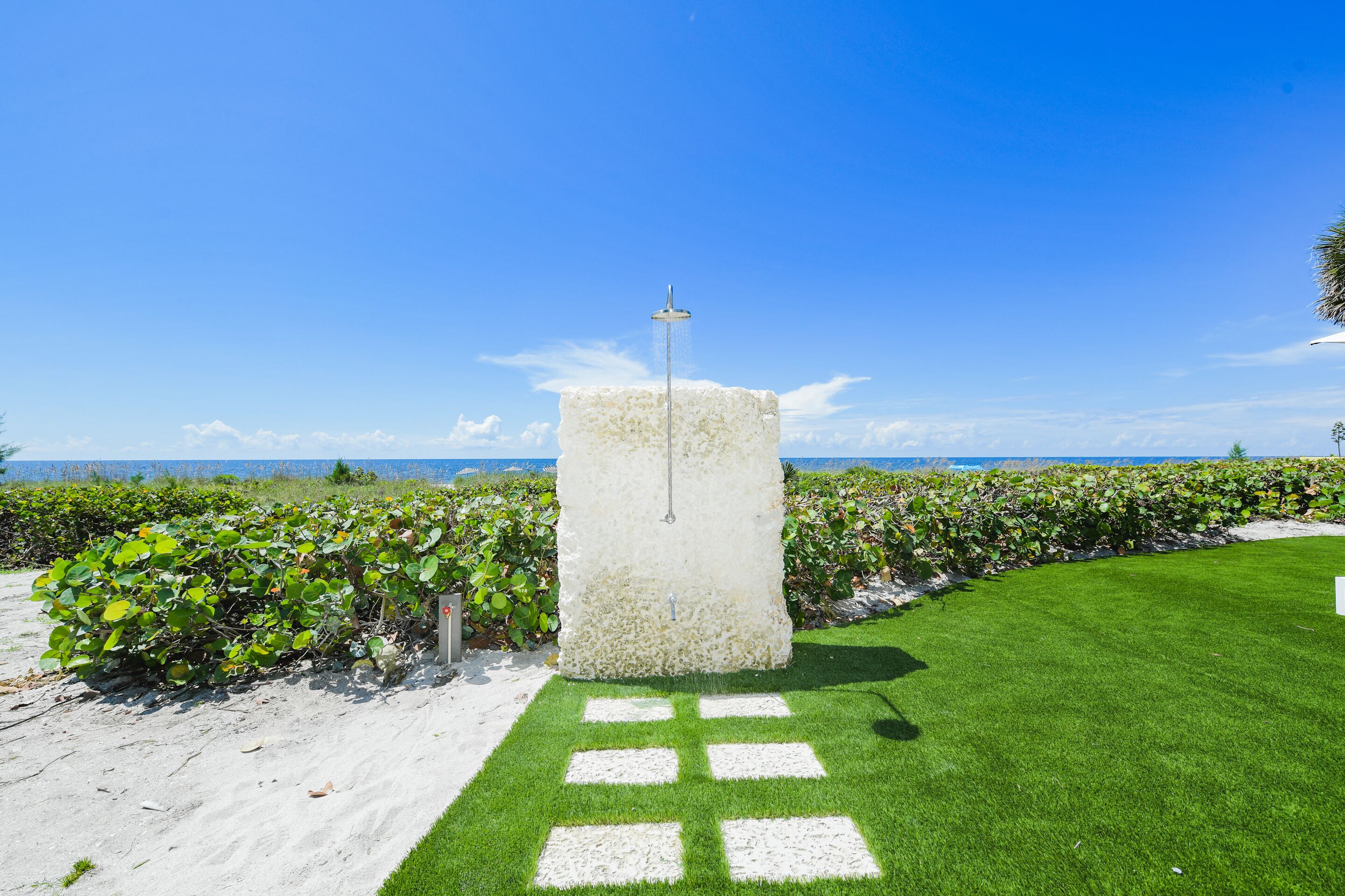 Une structure en béton surmontée d’une pomme de douche, entourée d’herbe verte et d’arbustes, avec un ciel bleu clair et l’océan en arrière-plan.