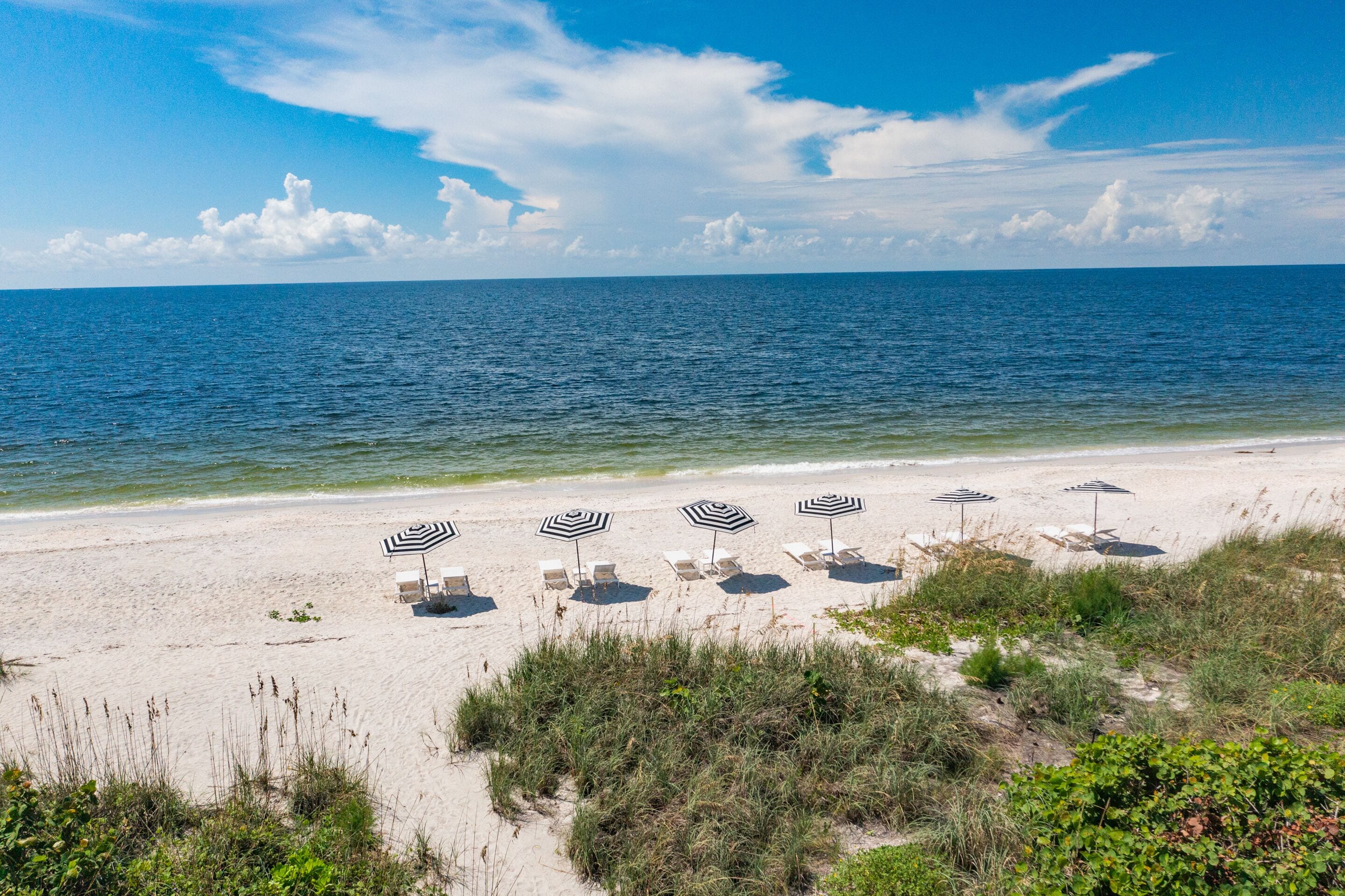 Ein Strand mit weißem Sand und klarem, blauem Meer. Mehrere Strandsonnenmatten und Sonnenschirme sind zum Sonnenbaden aufgestellt.
