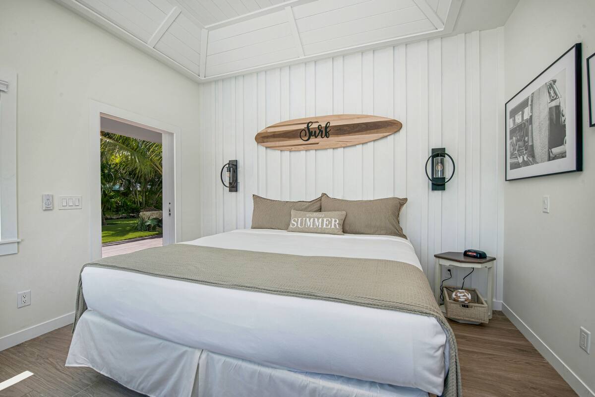 A bedroom with a bed, a wooden sign above the bed, a nightstand, and a view of a garden.