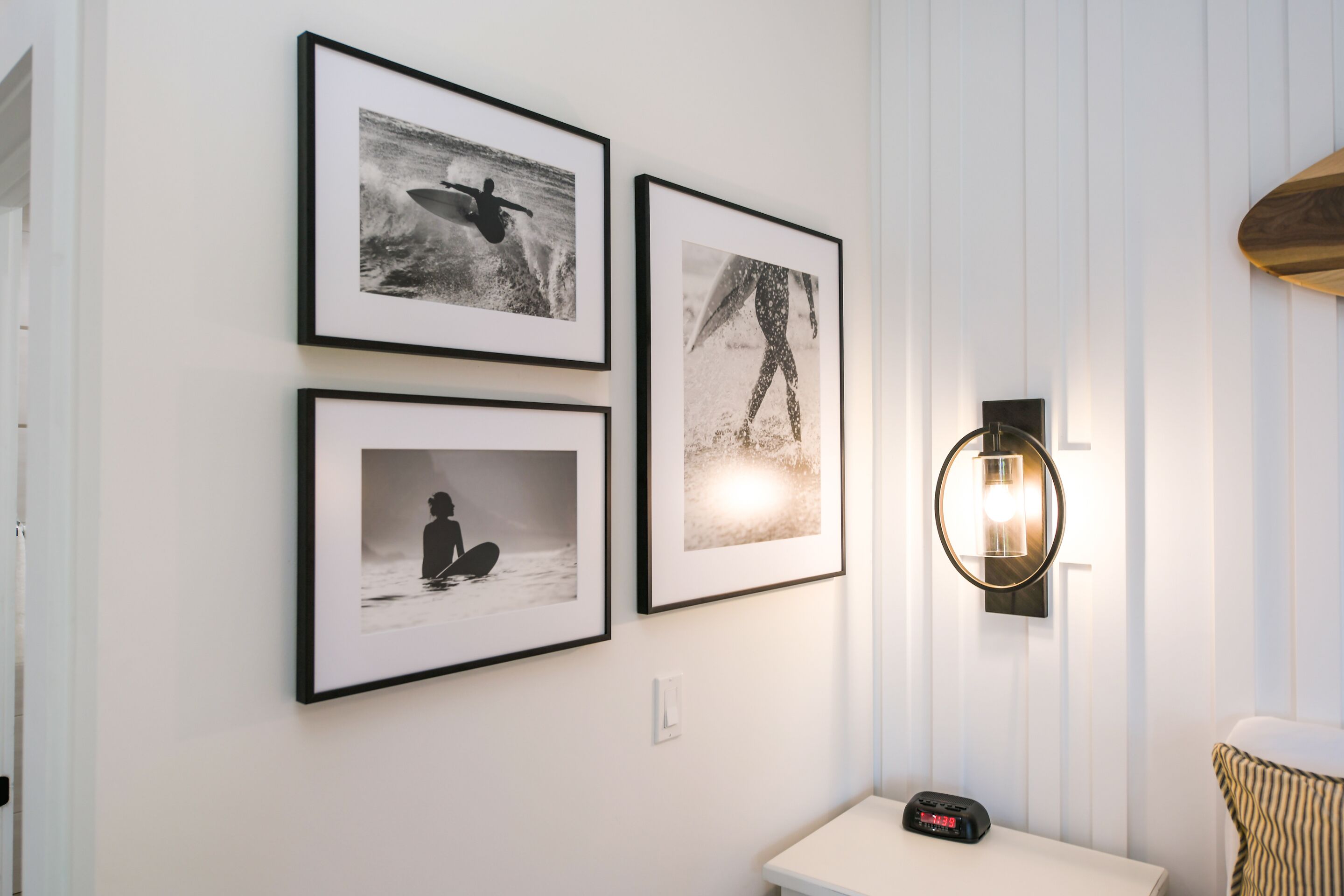 A wall with three framed photographs depicting surfing scenes, a small table with a clock, and a wall-mounted light fixture.