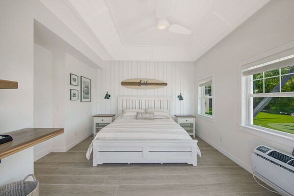 A bedroom with a bed, bedside tables, a ceiling fan, a window with a view, and a wall-mounted shelf.