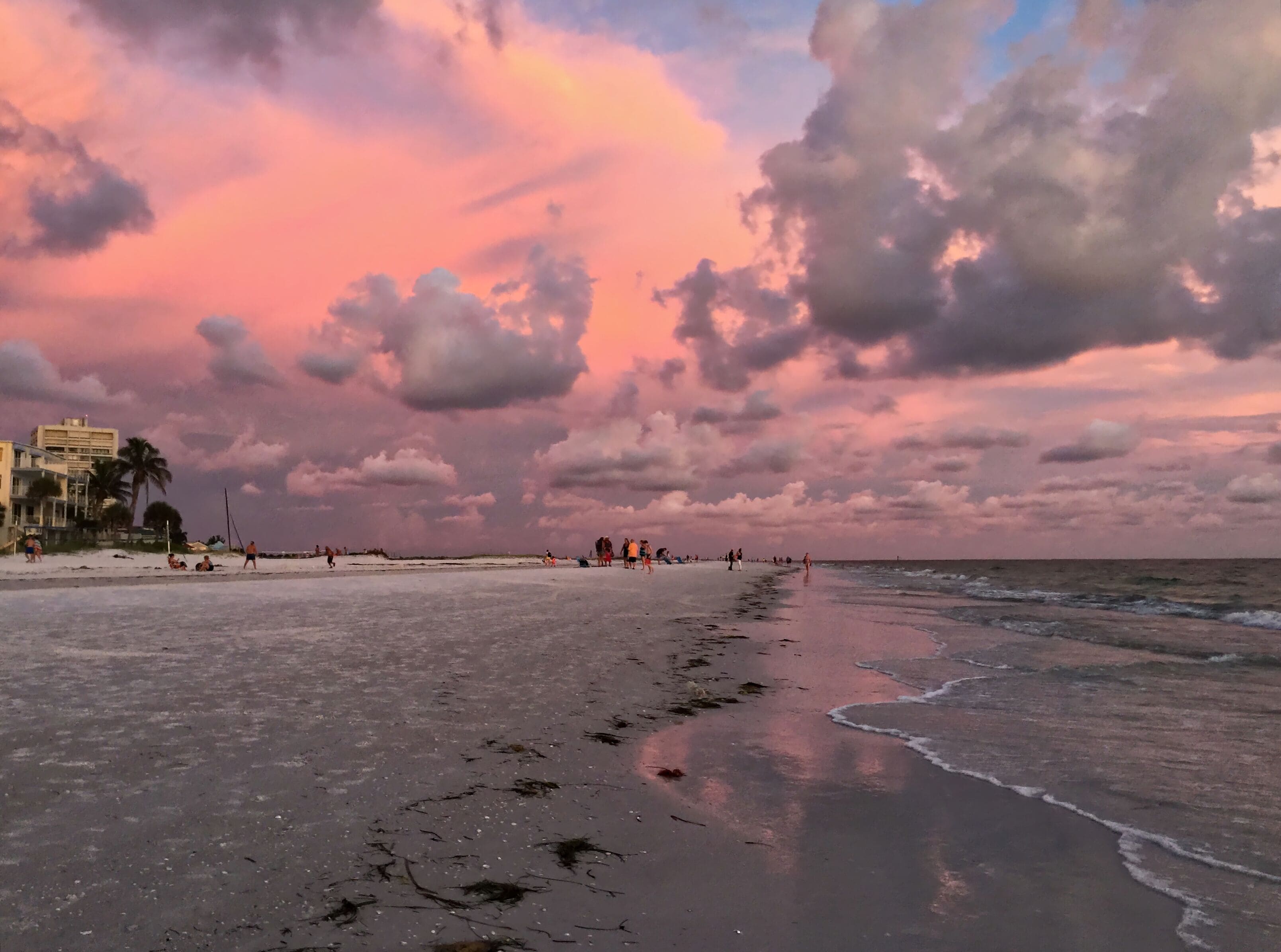 Une plage où l’on voit des gens se promener et s’asseoir, des bâtiments en arrière-plan et un ciel nuageux.
