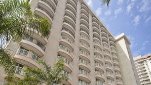 Un hotel de gran altura con varios balcones, una entrada moderna y palmeras en primer plano.
