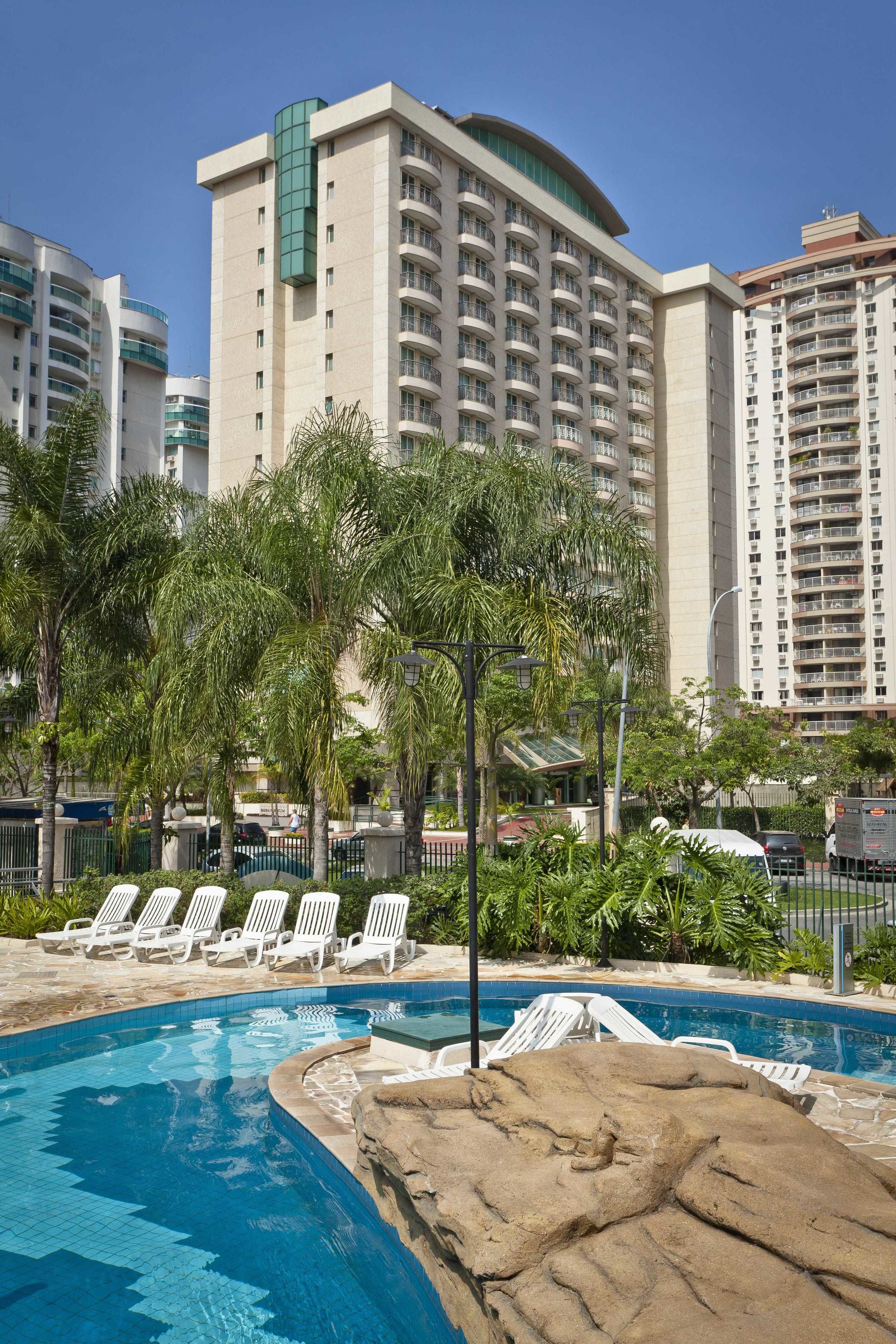 Zona de piscina de hotel con sillones reclinables, palmeras y un edificio grande al fondo.
