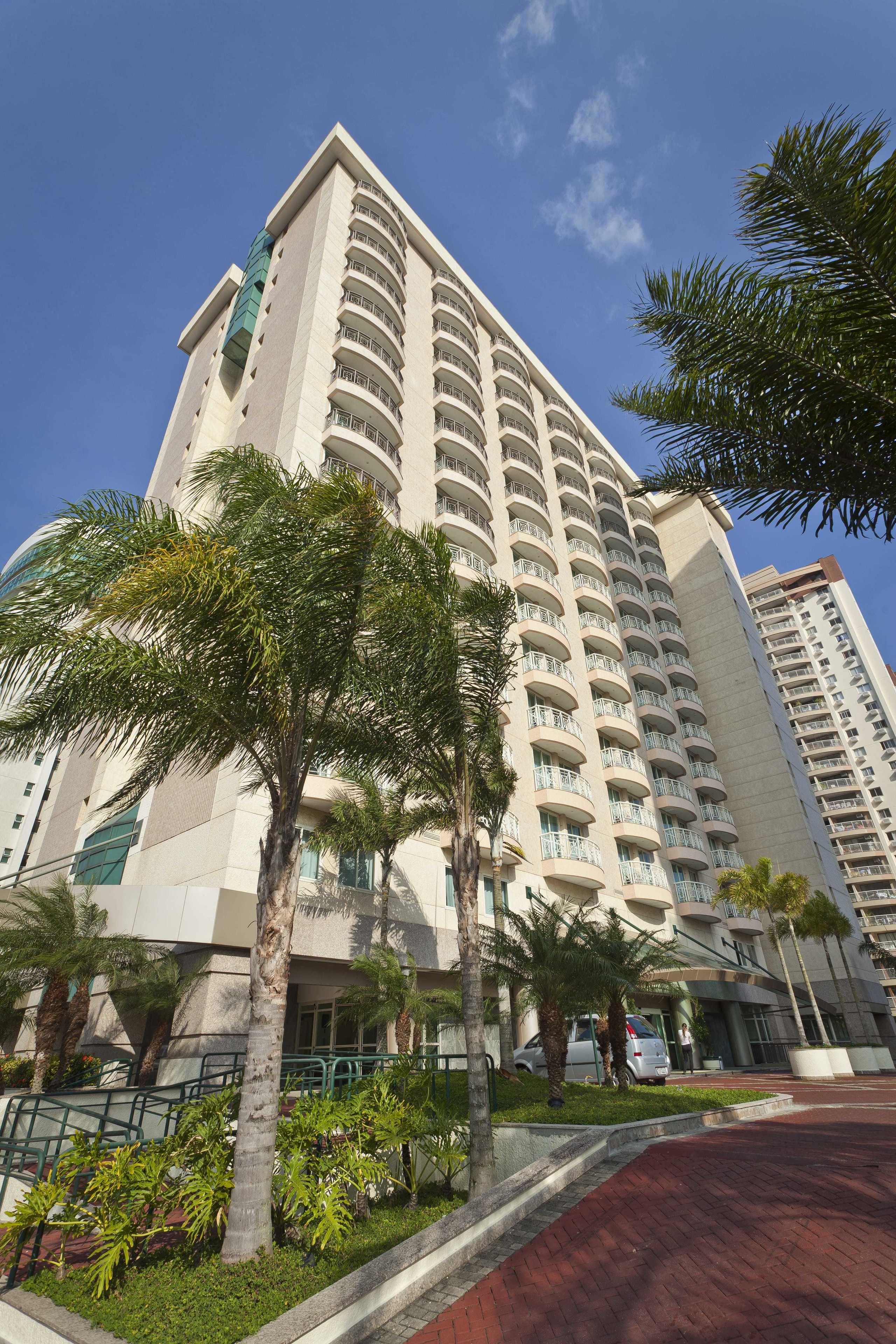 Hotel alto com várias varandas, rodeado por palmeiras e um caminho de tijolos vermelhos.