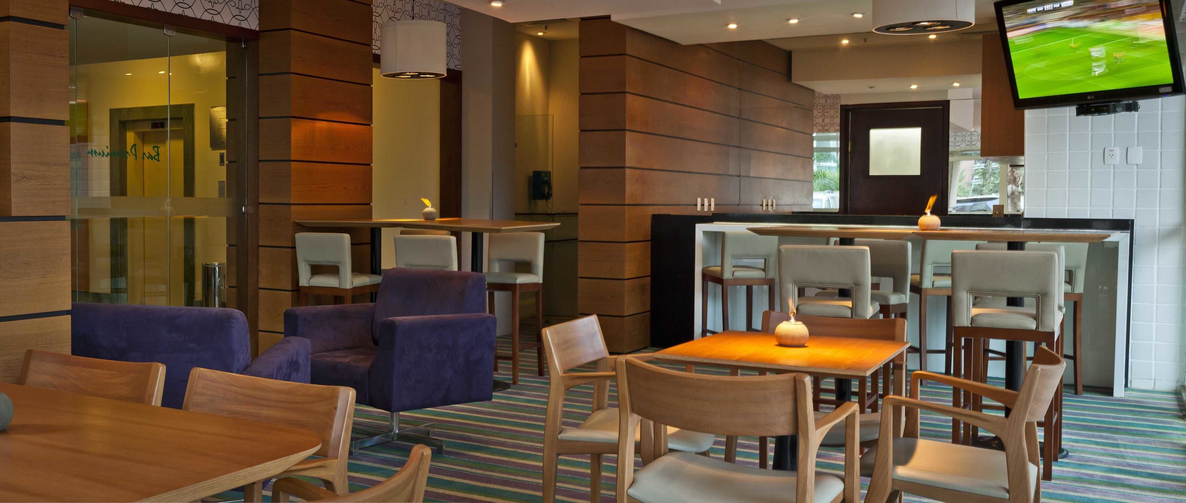 A modern hotel lobby with a bar area, seating arrangements, and a television displaying a sports event.