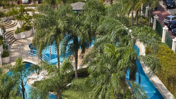 Una piscina grande y serpenteante rodeada de exuberantes palmeras y follaje, con un puente que conecta sus diferentes zonas.