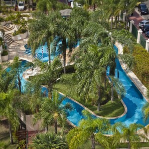 A large, winding pool surrounded by lush palm trees and greenery, with a bridge connecting different sections. - Bourbon Residence Barra da Tijuca | Rio de Janeiro (Rio de Janeiro)