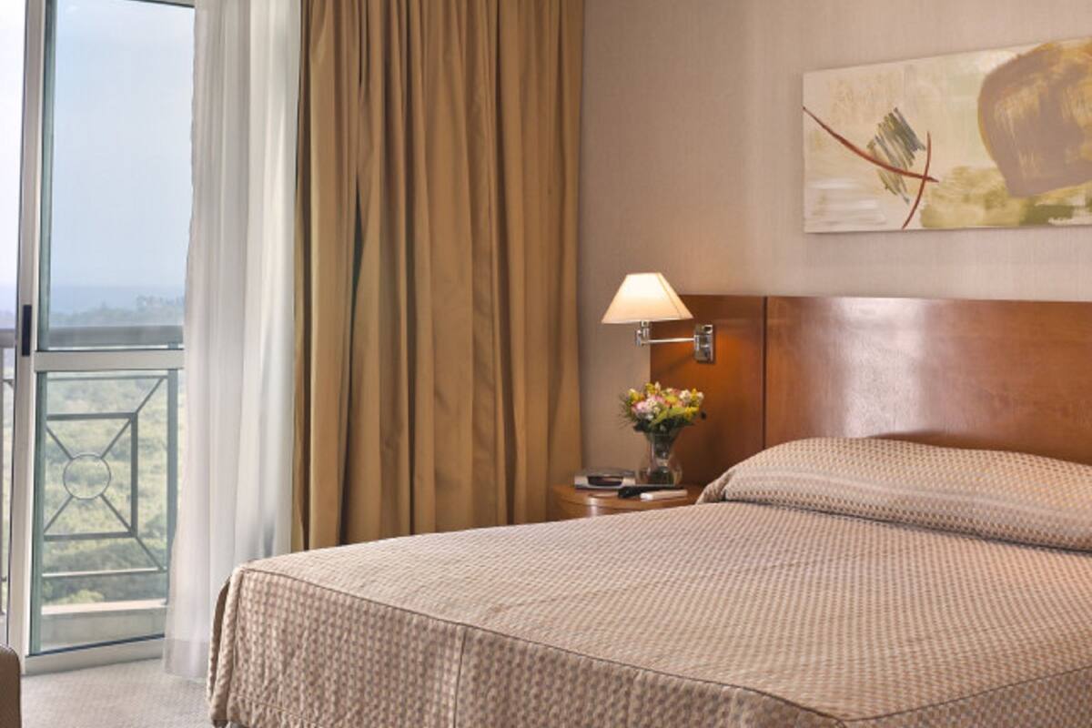 Ein Hotelzimmer mit einem großen Bett, einem Nachttisch mit Lampe, einem Bild an der Wand und Blick durch das Fenster.