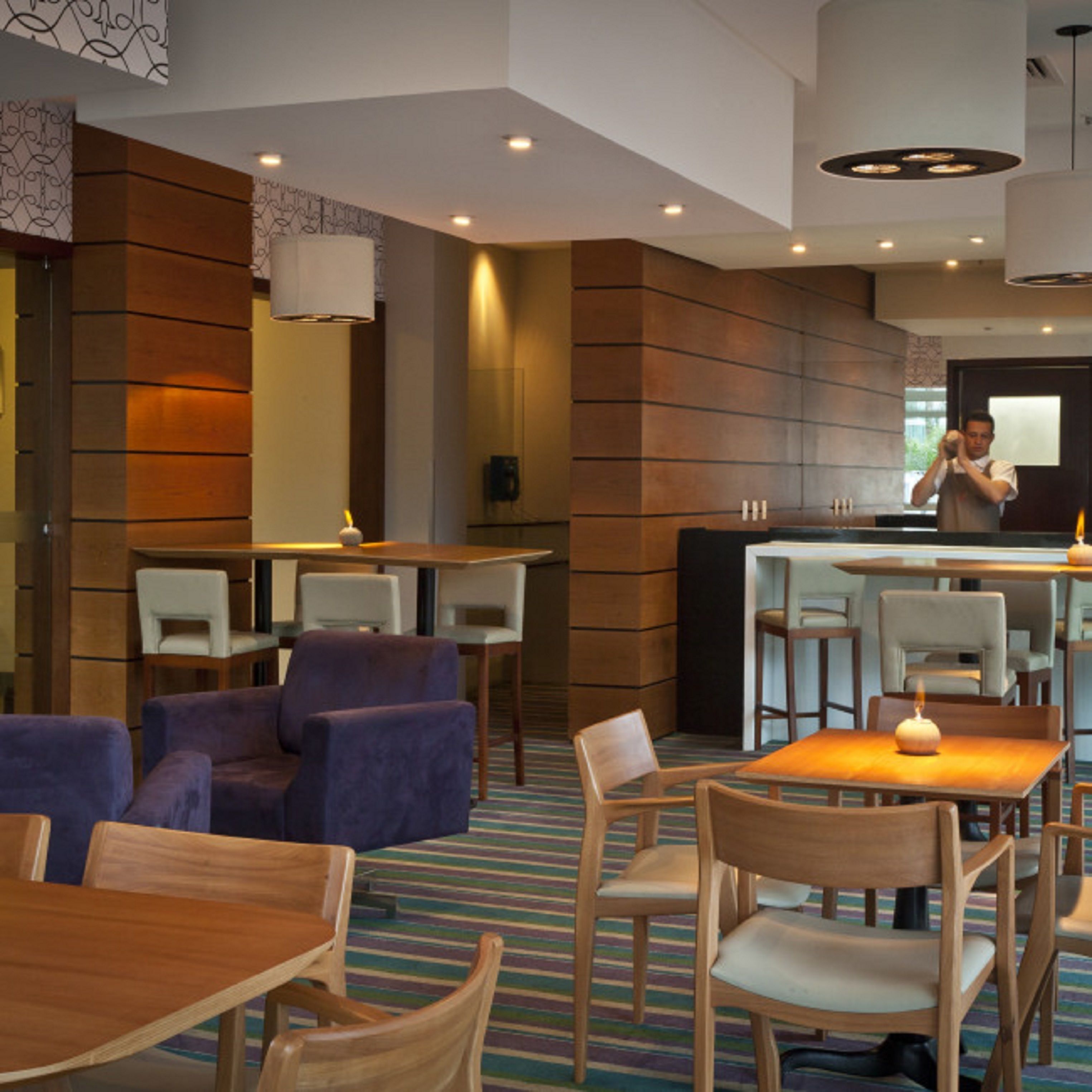 A modern hotel lobby with wooden panel walls, a central bar area, and seating arrangements including sofas and tables.