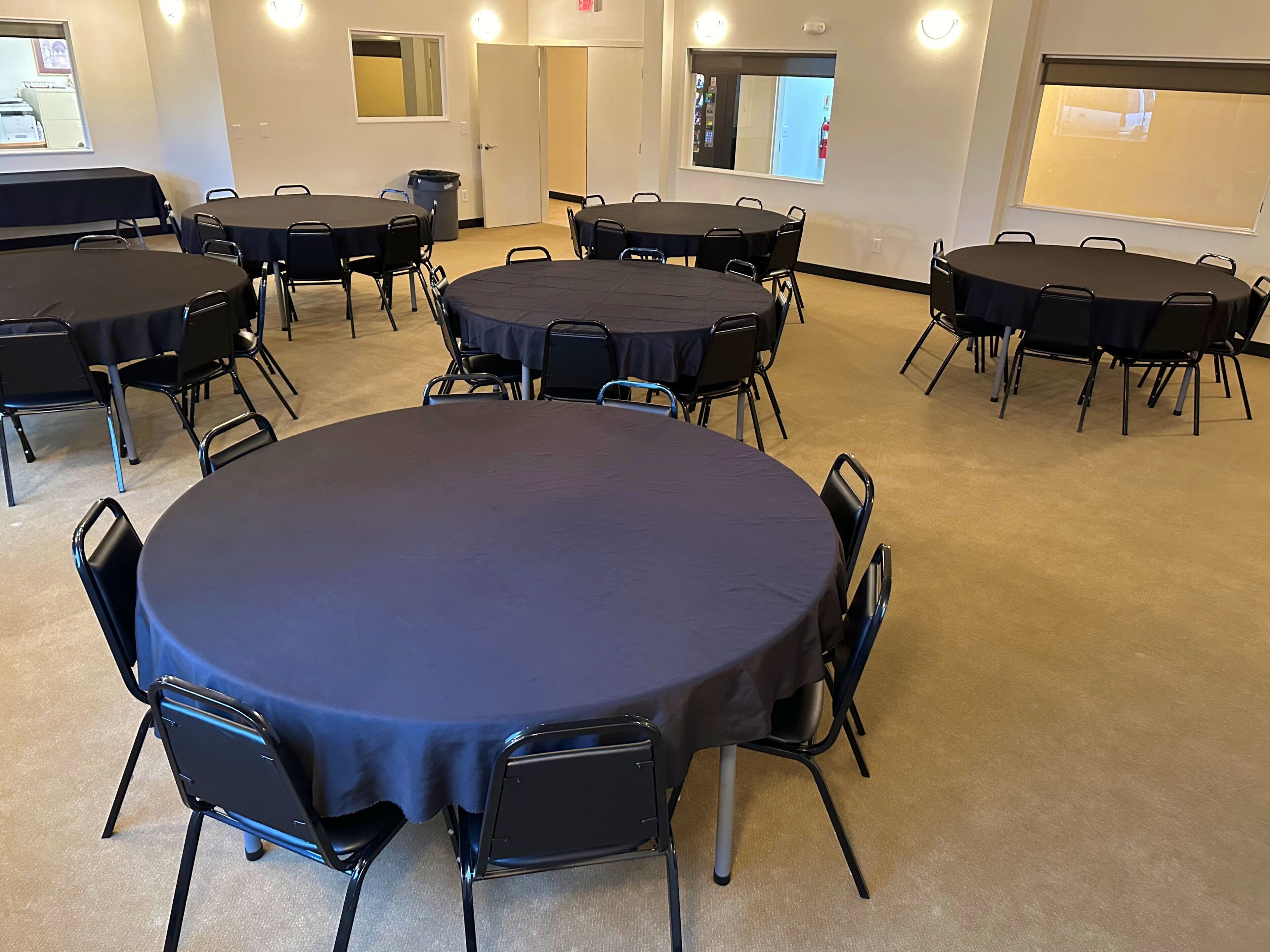 A conference room with multiple round tables and chairs, carpeted floor, and a large window with blinds.