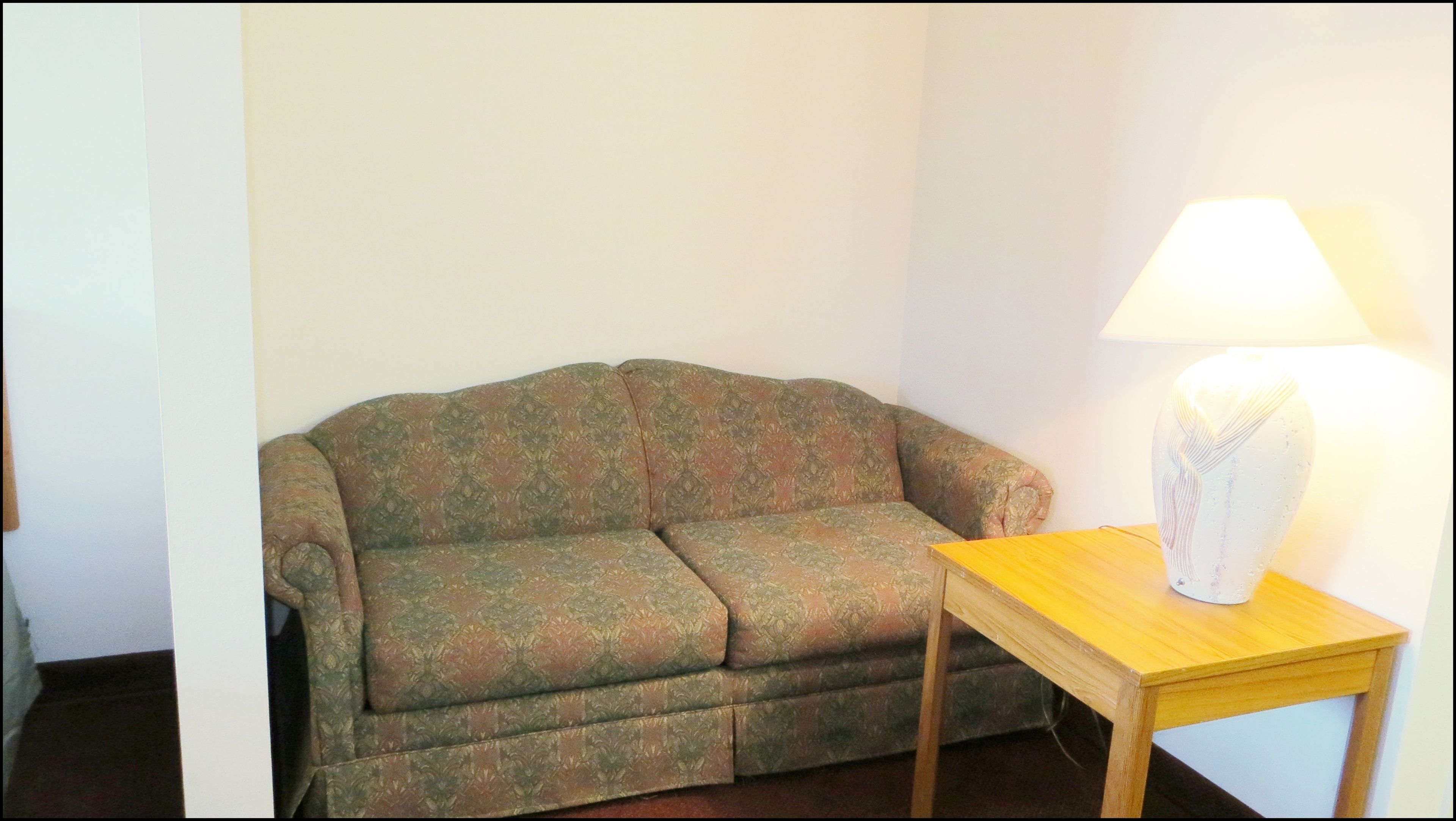 A small, single-seater sofa with a floral pattern, positioned against a plain wall. A wooden side table with a white lamp is placed in front of the sofa.