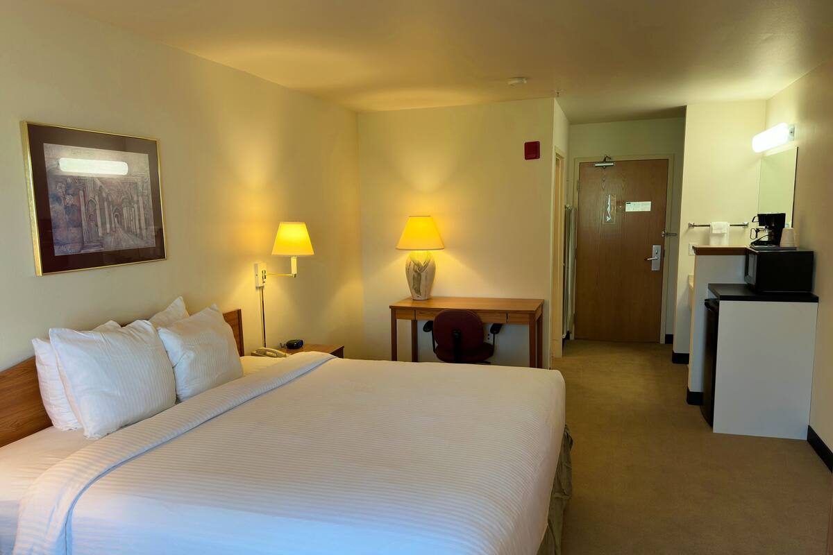 A hotel room with a large bed, two bedside lamps, a desk with a chair, a microwave, and a small table with a vase.