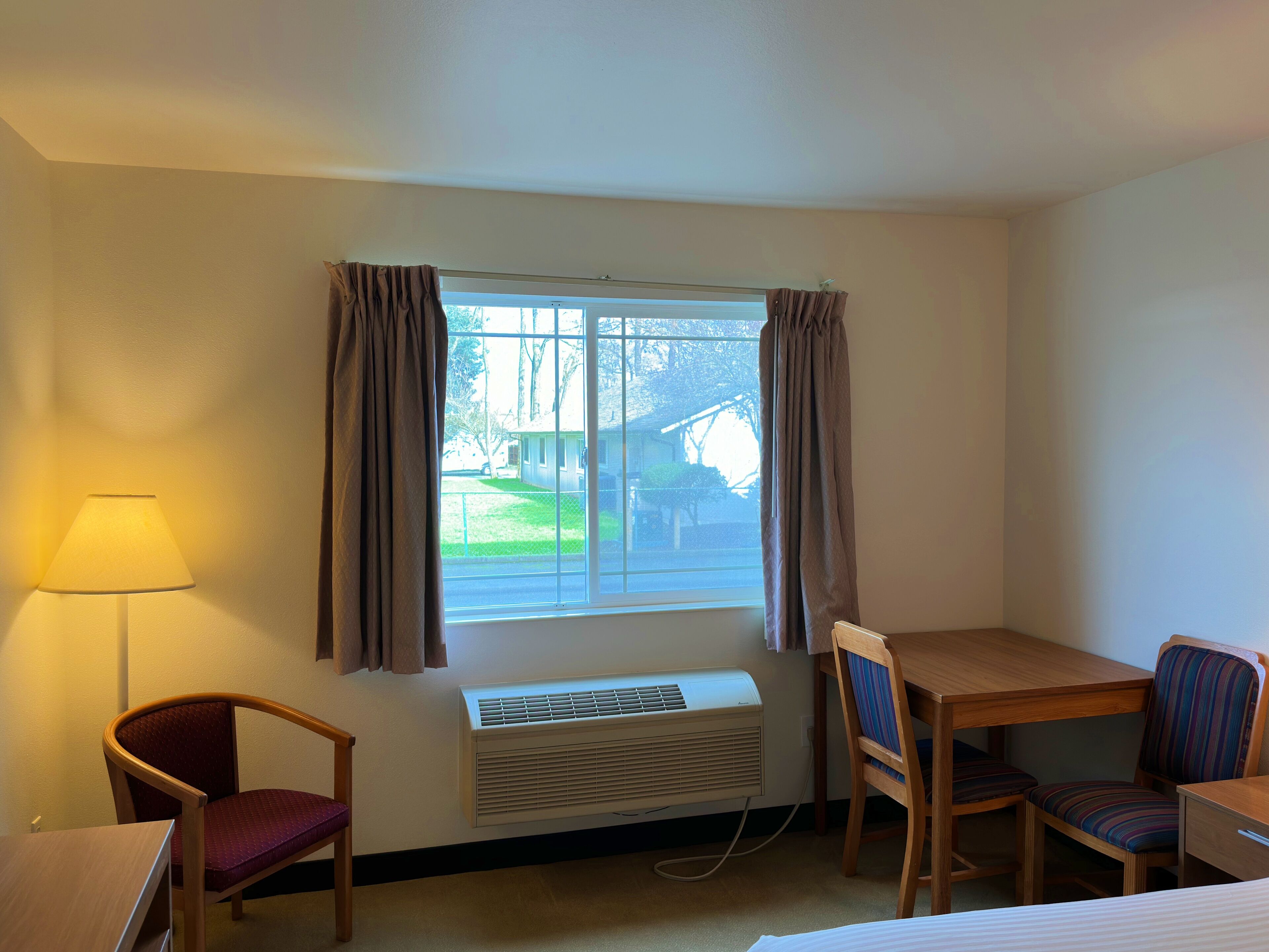A hotel room with a single bed, a wooden desk, two chairs, a lamp, an air conditioner, and a window with curtains.